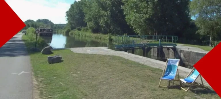 Paysage ensoleillé au bord d'un canal calme avec une écluse. Sur la rive herbeuse au premier plan, deux chiliennes bleues et blanches sont installées face à l'eau. Une voie verte longe le canal bordé d'arbres verdoyants, avec une péniche amarrée au loin.