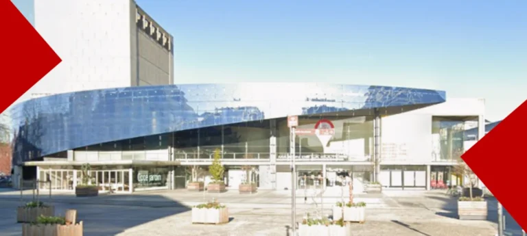 Gros plan de la façade vitrée incurvée de la Maison de la Culture d’Amiens (MCA), dont les reflets sombres bleu foncé masquent une partie de l'intérieur, prise depuis l'extérieur de jour. La structure est composée d'une haute tour blanche texturée sur la gauche et d'une longue section vitrée qui s'étend vers la droite. Dans le quart inférieur droit, le texte "CÔTÉ JARDIN MAISON DE LA CULTURE" est visible sur une vitre. L'image est encadrée par deux formes géométriques rouges : un triangle en haut à gauche et une flèche à droite, cachant partiellement le bâtiment.