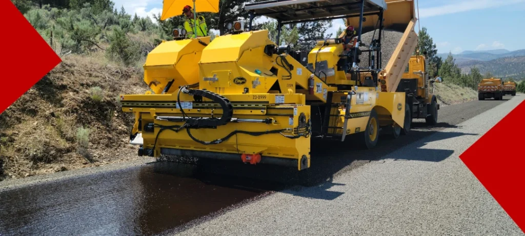 Gros plan d'une machine de revêtement routier jaune en action sur une route départementale dans l'Oise, appliquant de l'asphalte noir frais sur le bitume existant. Un travailleur en gilet de sécurité et un camion-benne sont visibles en arrière-plan sous un soleil éclatant.