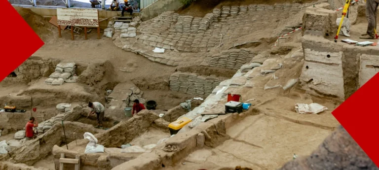 Vue plongeante d'un site de fouilles archéologiques en cours, avec plusieurs archéologues travaillant minutieusement au pinceau et à la truelle au milieu de structures en terre et de nombreux sacs de sable. Des équipements de fouille, une échelle et des panneaux d'information sont visibles. L'image est encadrée par deux coins rouges géométriques.