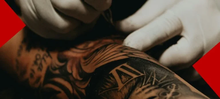 Gros plan détaillé de deux mains gantées de blanc travaillant avec précision sur un grand tatouage noir et gris complexe recouvrant un avant-bras. Le design présente des éléments de boussole et des chiffres romains. L'image est encadrée par deux formes géométriques rouges en haut à gauche et en bas à droite.