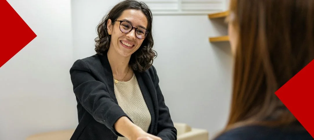 Portrait d'une femme professionnelle souriante, portant des lunettes et une veste noire sur un pull beige, en train de serrer la main d'une autre personne (hors champ) lors d'un entretien d'embauche. L'arrière-plan montre un bureau lumineux et moderne avec des étagères en bois. Des éléments graphiques rouges triangulaires encadrent les coins supérieurs gauches et inférieurs droits de l'image.