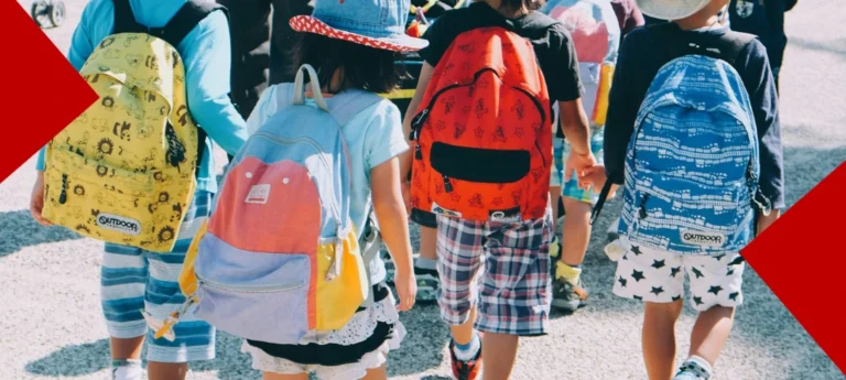 Vue de dos d'un groupe d'enfants marchant ensemble, portant chacun un sac à dos coloré et des vêtements estivaux (shorts, chapeaux de soleil). Les sacs à dos présentent divers motifs et couleurs vives comme le jaune, le rouge, le bleu et le pastel.