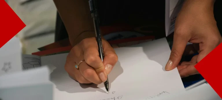 Vue rapprochée des mains d'une personne écrivant au stylo sur une feuille blanche. Elle porte une bague dorée ornée d'une pierre turquoise. Le cadre est agrémenté de deux triangles rouges vifs dans les coins opposés.