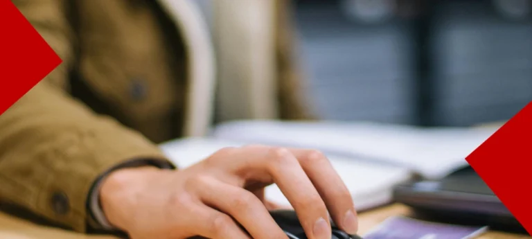 Gros plan sur la main d'une personne utilisant une souris d'ordinateur sur un bureau, avec un carnet ouvert en arrière-plan flou. Des formes géométriques rouges encadrent les coins de l'image.