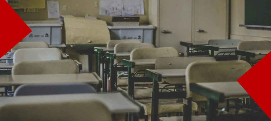 Une salle de classe vide avec plusieurs rangées de pupitres et de chaises en bois usés. L'arrière-plan montre des boîtes de rangement et des dessins affichés au mur. Deux formes géométriques rouges décorent les coins opposés de l'image.