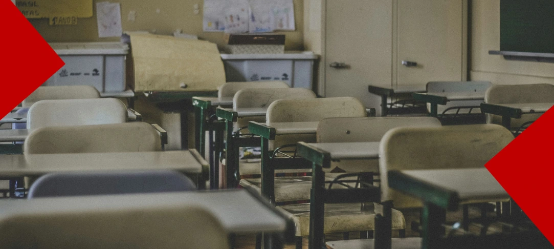 Une salle de classe vide avec plusieurs rangées de pupitres et de chaises en bois usés. L'arrière-plan montre des boîtes de rangement et des dessins affichés au mur. Deux formes géométriques rouges décorent les coins opposés de l'image.