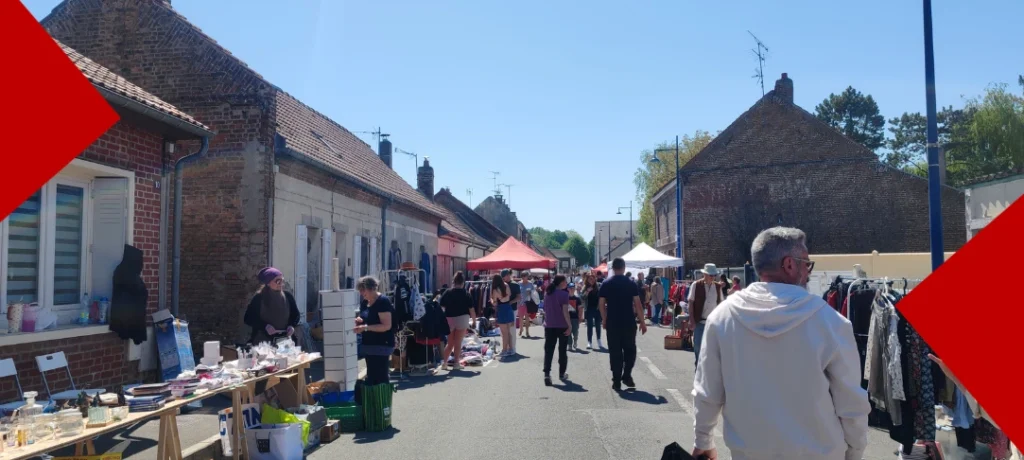Une rue ensoleillée bordée de maisons en briques rouges accueillant une brocante ou un vide-grenier. Des passants circulent entre les stands de vêtements et d'objets divers installés sur les trottoirs.