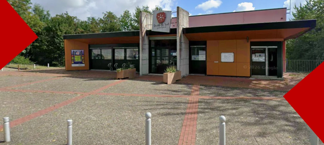 Vue extérieure d'un bâtiment de plain-pied, le centre culturel Étoile du Sud. La structure combine du béton gris, des boiseries et des surfaces orange. Au-dessus de la porte vitrée principale, un panneau affiche le nom du centre sous un blason rouge orné d'une étoile blanche. L'esplanade au premier plan est faite de pavés et de briques, avec quelques bacs à fleurs et des poteaux de signalisation gris. Des arbres verdoyants bordent le site sous un ciel légèrement nuageux.
