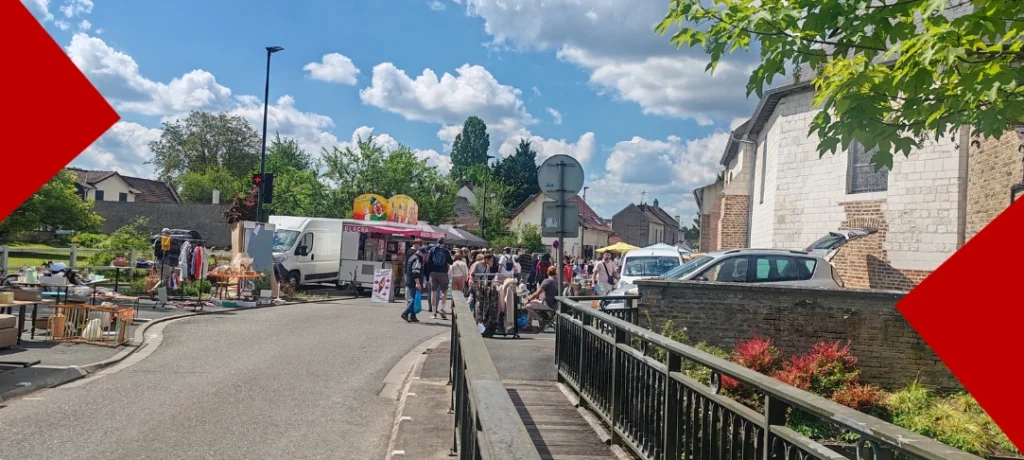 Photographie d'un vide-greniers en plein air sous un ciel bleu parsemé de nuages blancs. À gauche, des étals présentent des vêtements et du mobilier. Au centre, un camion de glaces blanc stationne près d'une foule de visiteurs. Sur la droite, une église traditionnelle en pierre blanche borde la rue. Un garde-corps métallique au premier plan accentue la perspective de la scène de village.