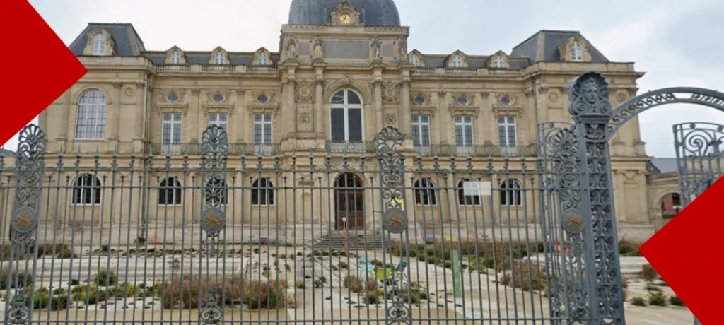 Une photo du bâtiment néo-classique du Musée de Picardie en pierre d'Amiens, avec une façade ornée et un toit mansardé, prise derrière une grille en fer forgé décorative. Des éléments géométriques rouges sont visibles dans les coins supérieur gauche et inférieur droit.