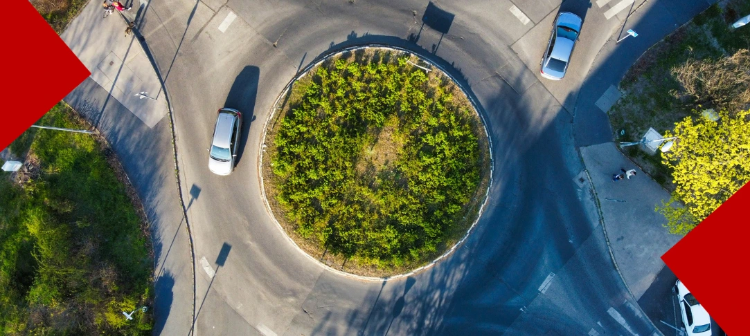 Vue aérienne en plongée d'un rond-point arboré avec deux voitures en circulation, encadrée par deux formes géométriques rouges dans les angles opposés.