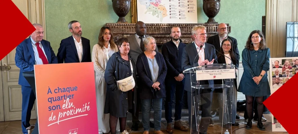 Le maire d’Amiens, Frédéric Fauvet, s’exprime derrière un pupitre lors d’une conférence de presse sur la réforme de la démocratie locale, entouré de plusieurs élus référents. À l'avant-plan gauche, un panneau d'affichage coloré affiche le slogan « À chaque quartier son élu de proximité ». En arrière-plan, une carte des nouveaux quartiers d'Amiens est visible au-dessus de la cheminée