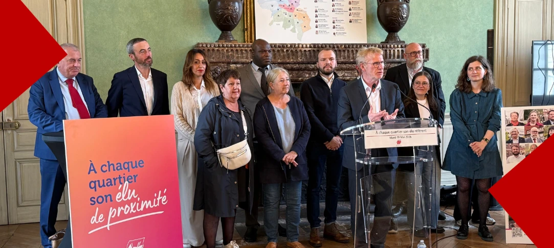 Le maire d’Amiens, Frédéric Fauvet, s’exprime derrière un pupitre lors d’une conférence de presse sur la réforme de la démocratie locale, entouré de plusieurs élus référents. À l'avant-plan gauche, un panneau d'affichage coloré affiche le slogan « À chaque quartier son élu de proximité ». En arrière-plan, une carte des nouveaux quartiers d'Amiens est visible au-dessus de la cheminée