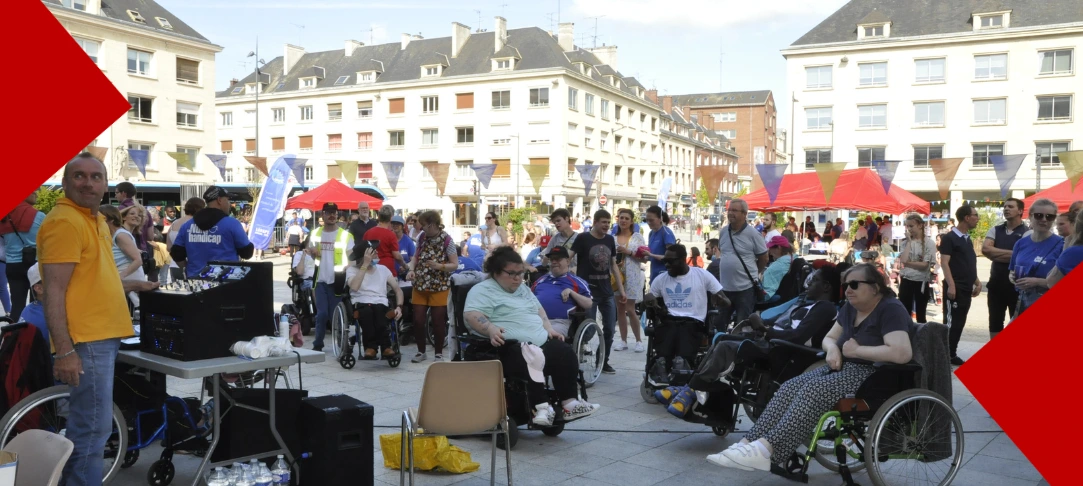 Une vue large d'un événement en plein air sur une place de ville pavée, entourée d'immeubles résidentiels clairs. De nombreuses personnes sont rassemblées, dont plusieurs sont en fauteuil roulant (manuel ou électrique). Au premier plan à gauche, un homme en t-shirt jaune se tient près d'une table avec une régie de sonorisation et des bouteilles d'eau. Au centre et à droite, les participants discutent et sourient. En arrière-plan, on aperçoit des tentes rouges et des guirlandes de fanions colorés suspendues, renforçant l'aspect festif de l'événement. Le visuel est encadré par deux grands triangles rouges texturés dans les coins supérieur gauche et inférieur droit.