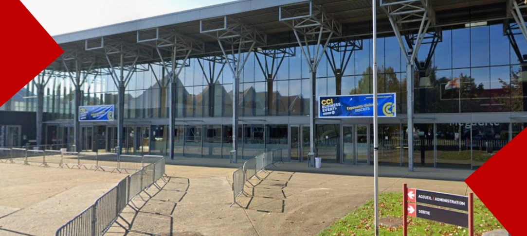 Une vue grand angle de l'entrée principale en verre de l'espace événementiel Mégacité à Amiens. La façade est surmontée d'une structure de toiture en treillis métallique. L'avant-plan est une zone d'attente pavée entourée de barrières métalliques de contrôle des foules. Sur la façade vitrée, une bannière bleue et jaune indique « CCI BUSINESS EVENT » et « Exposants - Visiteurs INSCRITS ». À droite, un panneau d'orientation bilingue pointe vers « ACCUEIL / ADMINISTRATION » et « SORTIE ». Des éléments de design en forme de coins rouges encadrent les coins supérieur gauche et inférieur droit
