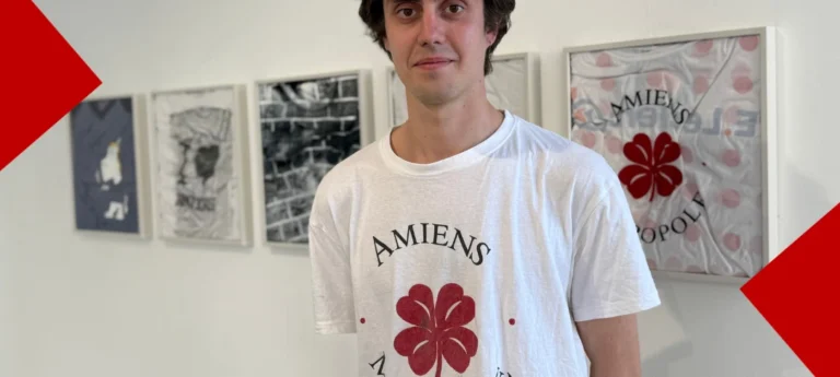 Portrait à mi-corps de l'artiste Martin Gobert, un jeune homme brun portant un t-shirt blanc de sa création où est inscrit "Amiens" au-dessus d'un trèfle à quatre feuilles rouge. En arrière-plan, sur un mur blanc, plusieurs de ses œuvres sont exposées et encadrées, incluant un autre t-shirt sérigraphié similaire présenté comme un maillot de collection. Des formes géométriques rouges encadrent les coins supérieurs gauche et inférieurs droit de l'image.