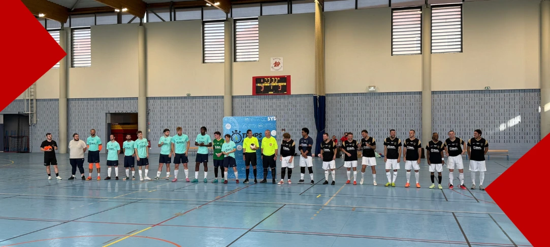 Une rangée de joueurs de futsal et d'arbitres alignés sur un terrain intérieur en résine bleue, juste avant le début d'un match. Au centre, deux arbitres en maillot jaune fluo se tiennent devant un panneau publicitaire bleu. À leur gauche, une équipe porte des maillots turquoise et des shorts noirs, tandis qu'à leur droite, l'autre équipe porte des maillots noirs avec des détails dorés et des shorts blancs. En arrière-plan, le mur de la salle de sport comporte un tableau d'affichage électronique rouge et de hautes fenêtres rectangulaires. Les coins supérieur gauche et inférieur droit de l'image sont recouverts par de grands triangles graphiques rouges.
