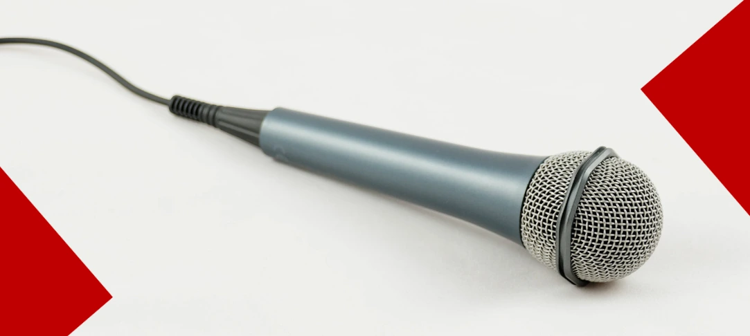 Un microphone dynamique gris métallique avec une grille de protection argentée, posé sur un fond blanc épuré. Un câble noir part du manche vers la gauche. Deux grands triangles décoratifs de couleur rouge vif encadrent l'image dans le coin inférieur gauche et le coin supérieur droit.