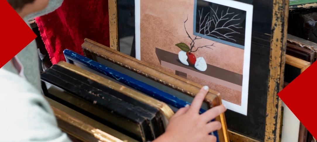 Gros plan sur une main de femme tenant un cadre photo en bois doré dans une brocante ou un marché aux puces, examinant une œuvre d'art surréaliste représentant une pomme rouge cassée en deux et une feuille verte reposant sur une surface plate, avec des branches d'arbre nues s'étendant à partir de la pomme. L'image est encadrée par deux formes géométriques rouges en haut à gauche et en bas à droite.