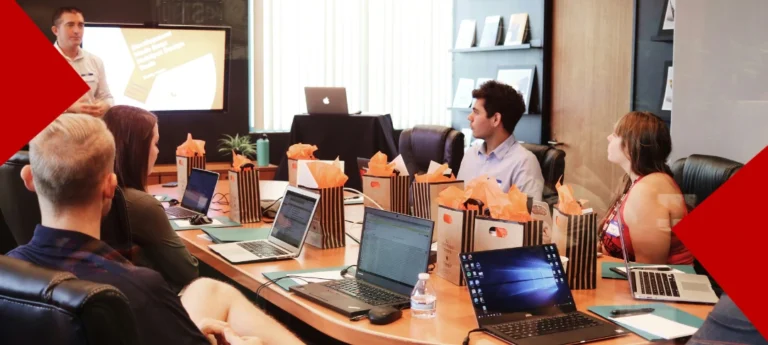 Des personnes sont assises autour d'une table, des ordinateurs portables devant les yeux. Le formateur est debout devant un écran.