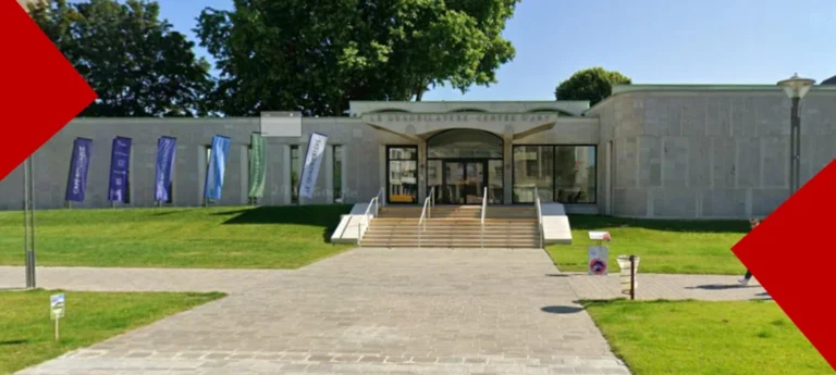 Façade extérieure du centre d'art Le Quadrilatère à Beauvais, un bâtiment moderne en pierre claire avec une large entrée vitrée et un escalier central. Des drapeaux colorés sont alignés devant la pelouse sur la gauche et une allée pavée mène à l'entrée. L'image est encadrée par deux triangles rouges dynamiques dans les coins.