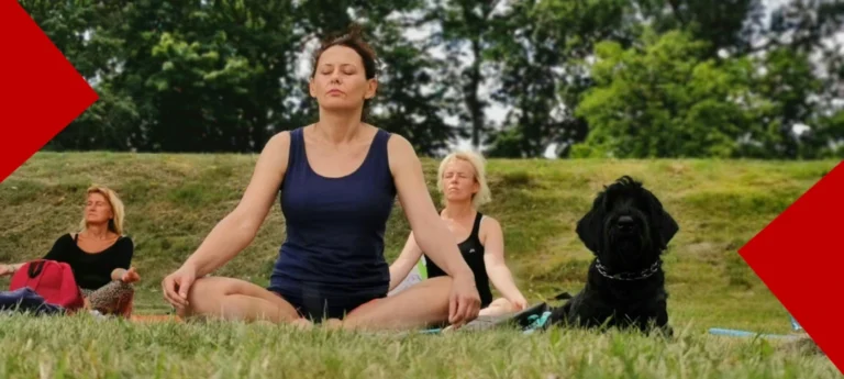 Une femme en débardeur bleu pratique la méditation assise en tailleur sur l'herbe d'un parc, les yeux fermés. Derrière elle, deux autres femmes sont également en méditation et un chien noir est assis calmement à sa droite. L'image dégage une ambiance sereine et de bien-être en plein air, encadrée par des coins rouges géométriques sur les bords.