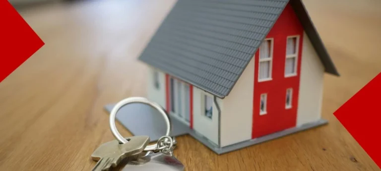 Une petite maison miniature blanche avec un toit gris et une façade rouge posée sur une table en bois clair. Au premier plan, un trousseau de clés avec un porte-clés en forme de maison en métal argenté est posé à côté de la maquette, symbolisant l'accession à la propriété ou un projet immobilier abouti.