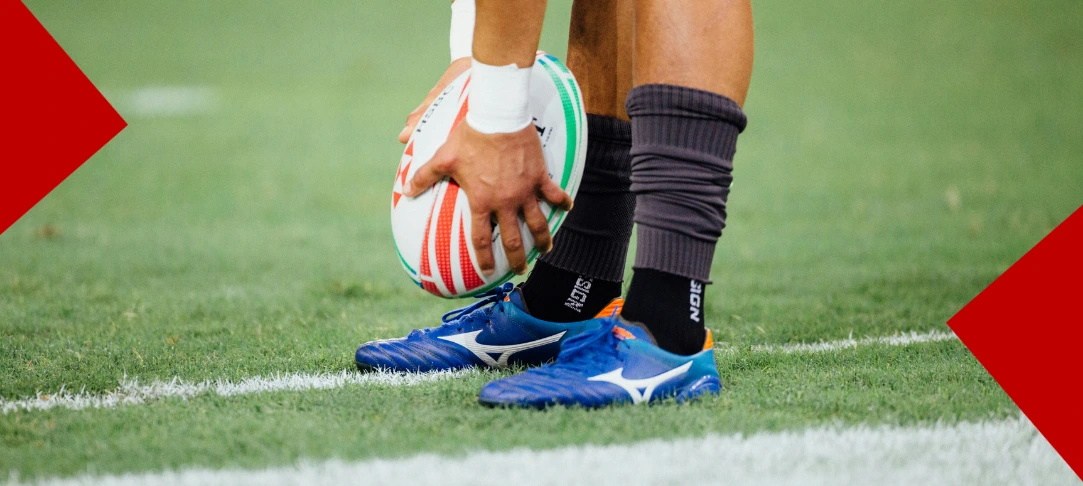 Gros plan sur les pieds d'un joueur de rugby en crampons bleus Mizuno, les mains gantées de blanc positionnant un ballon de rugby blanc et rouge sur la pelouse marquée d'une ligne blanche.