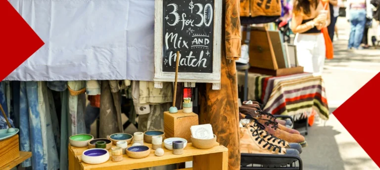 Vue rapprochée d'un stand de marché artisanal en extérieur présentant des poteries colorées, des vêtements vintage et des chaussures sur des étals en bois, avec une pancarte à la craie indiquant une offre promotionnelle.