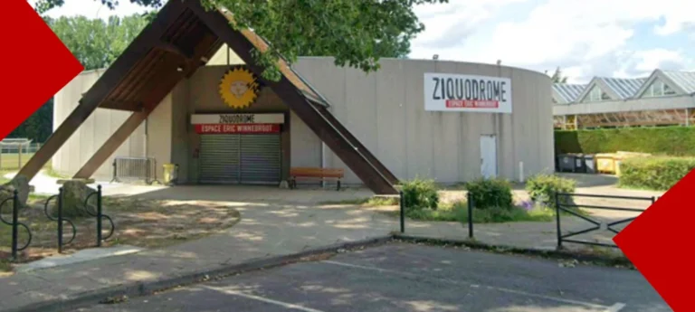 Photographie de l'entrée du Ziquodrome, sous-titré "Espace Éric Winnebroot". Le bâtiment présente une structure circulaire en béton gris. L'entrée principale est abritée par une grande structure en bois en forme de V inversé, surmontée d'un logo représentant un soleil jaune. À droite, un grand panneau affiche le nom du lieu. Le premier plan montre un parking et un petit espace vert, tandis que des triangles rouges habillent les coins supérieur gauche et inférieur droit de l'image.