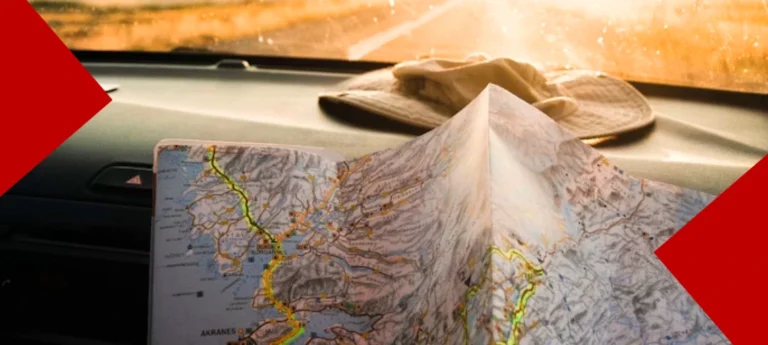 Vue de l'intérieur d'une voiture avec une carte routière ouverte sur le tableau de bord et un chapeau beige, face à une route ensoleillée.