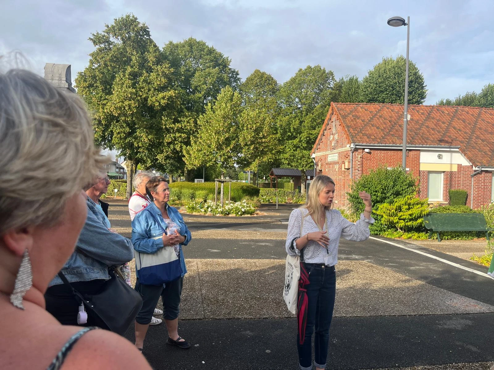 En extérieur, une jeune femme blonde vêtue d'un haut blanc et d'un jean parle en gesticulant de la main à un groupe de personnes qui l'écoutent. À l'arrière-plan se trouvent des arbres verts et une maison traditionnelle en briques rouges sous un ciel léger.